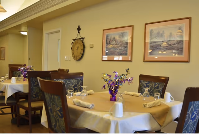A dining area with round tables covered in beige tablecloths and set with napkins, glasses, and white mugs. Each table has a small purple vase with colorful artificial flowers. The room has wooden chairs with patterned cushions, light yellow walls, two framed landscape paintings, and a large decorative clock on the wall.