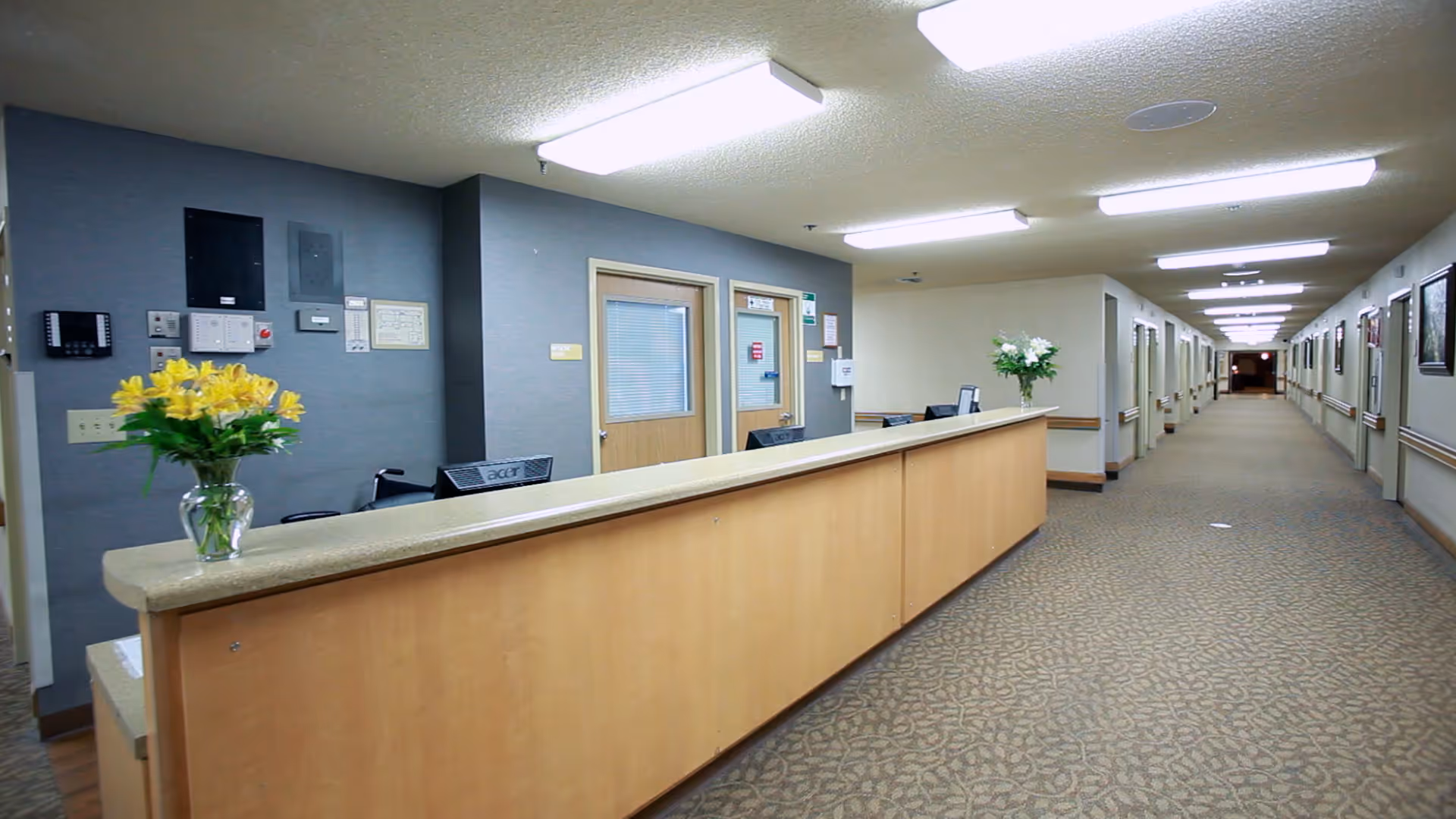 Interior view of a senior living facility hallway with a long wooden reception desk on the left side, decorated with a vase of yellow flowers. The hallway is carpeted with patterned flooring and has multiple doors along both sides. The ceiling has fluorescent lighting panels.