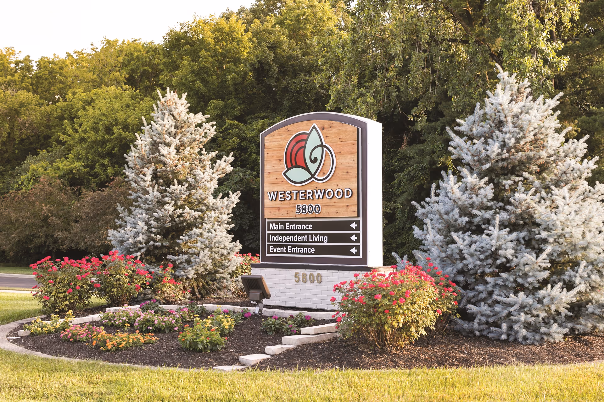 Outdoor landscaped area with a sign for Westerwood at 5800, surrounded by flowering plants and evergreen trees, with directions to Main Entrance, Independent Living, and Event Entrance.