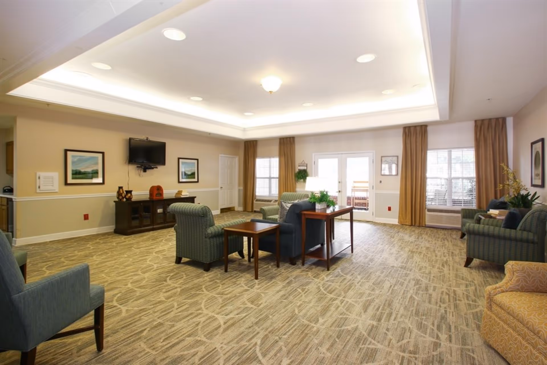 A spacious and well-lit living room area in a senior living community with several upholstered armchairs and sofas arranged around wooden side tables. The room features a patterned carpet, beige walls with framed artwork, a wall-mounted flat-screen TV, large windows with brown curtains, and a glass door leading outside. The ceiling has recessed lighting and a central light fixture.
