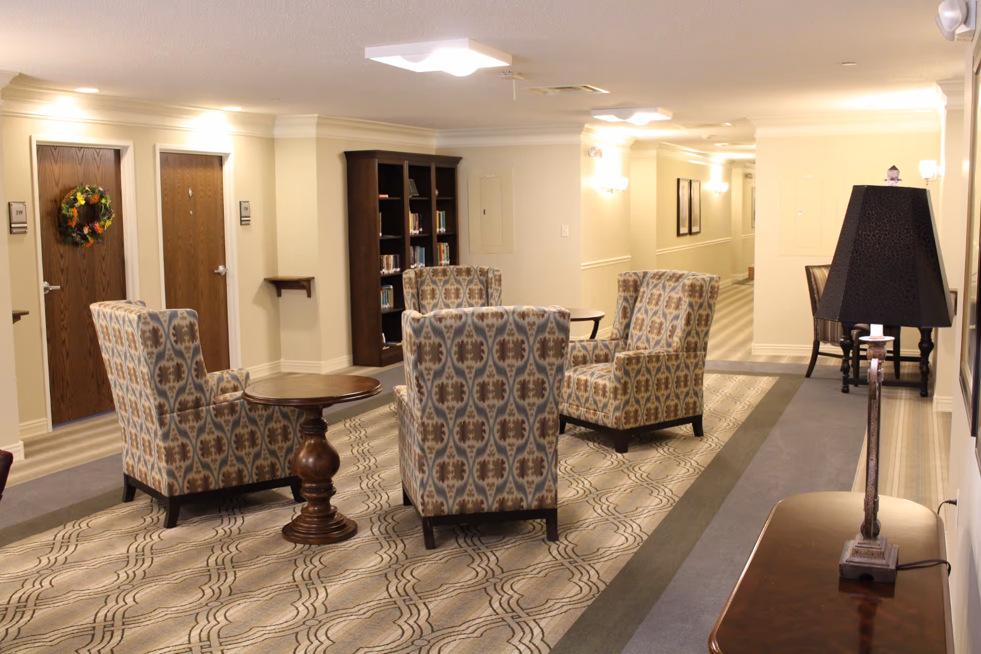 A cozy sitting area in a hallway of an assisted living facility with four patterned armchairs arranged around a small round wooden table. There is a bookshelf filled with books against the wall, two wooden doors with room numbers and a decorative wreath on one door, and a hallway extending into the background. A table with a lamp is visible on the right side.
