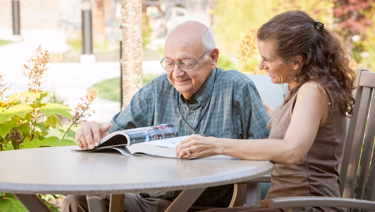 An elderly man and a woman sitting at an outdoor table, smiling and looking at a book together. They are surrounded by greenery and plants in a garden-like setting.