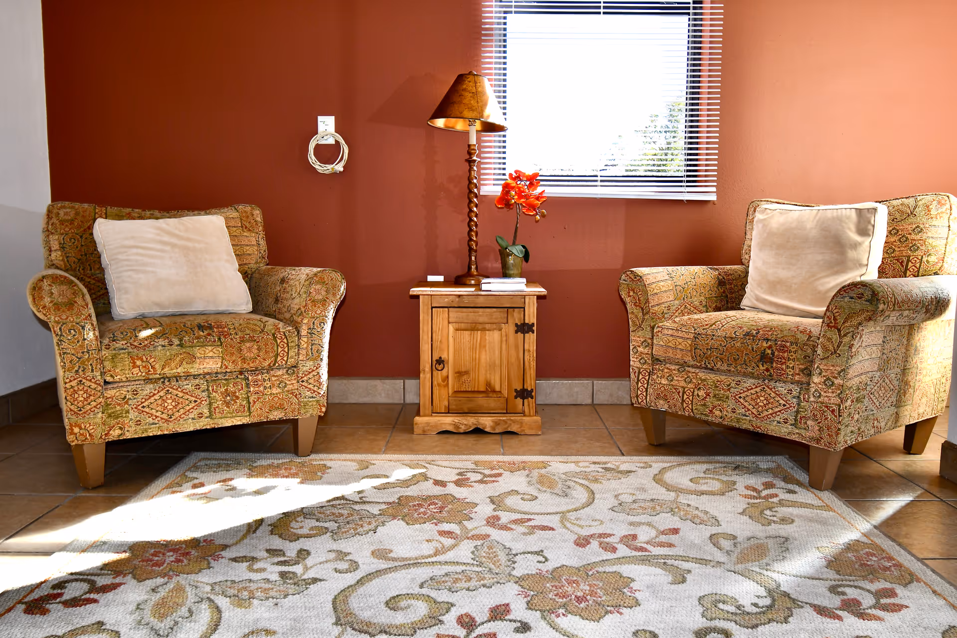 Two patterned armchairs with white cushions facing each other with a small wooden side table in between. The table holds a lamp and a small plant with red flowers. Behind the table is a window with blinds partially open, and the floor is tiled with a floral patterned rug in front of the chairs.
