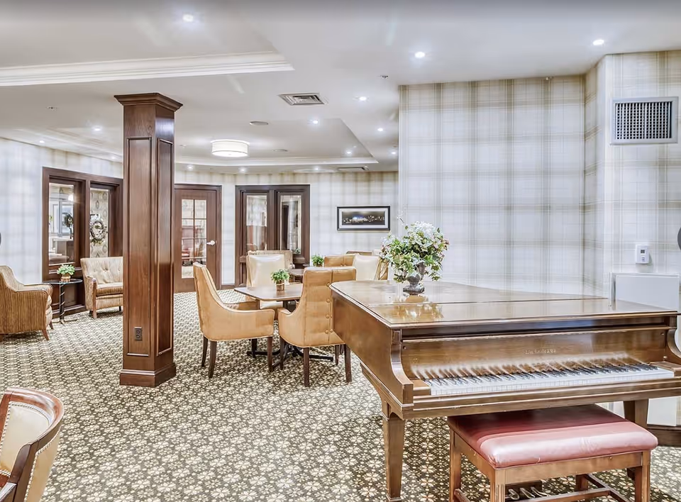 A well-lit interior common area with a polished wooden grand piano and a matching bench in the foreground. The room features patterned carpet flooring, beige upholstered chairs around small tables, and a wooden pillar. The walls have a light plaid wallpaper, and there are glass-paneled doors and framed artwork on the walls.