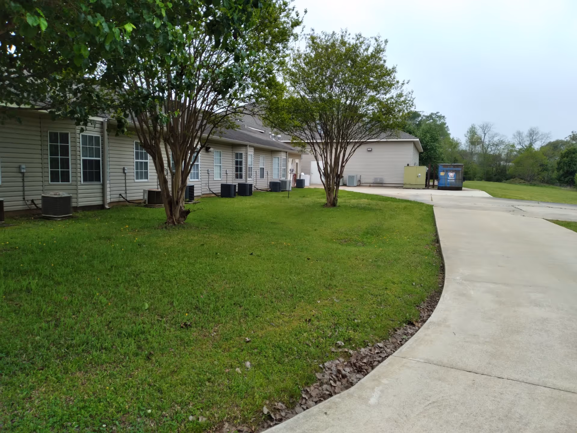 Exterior side view of a single-story assisted living building with a grassy lawn, trees, windows, and a curved concrete driveway.