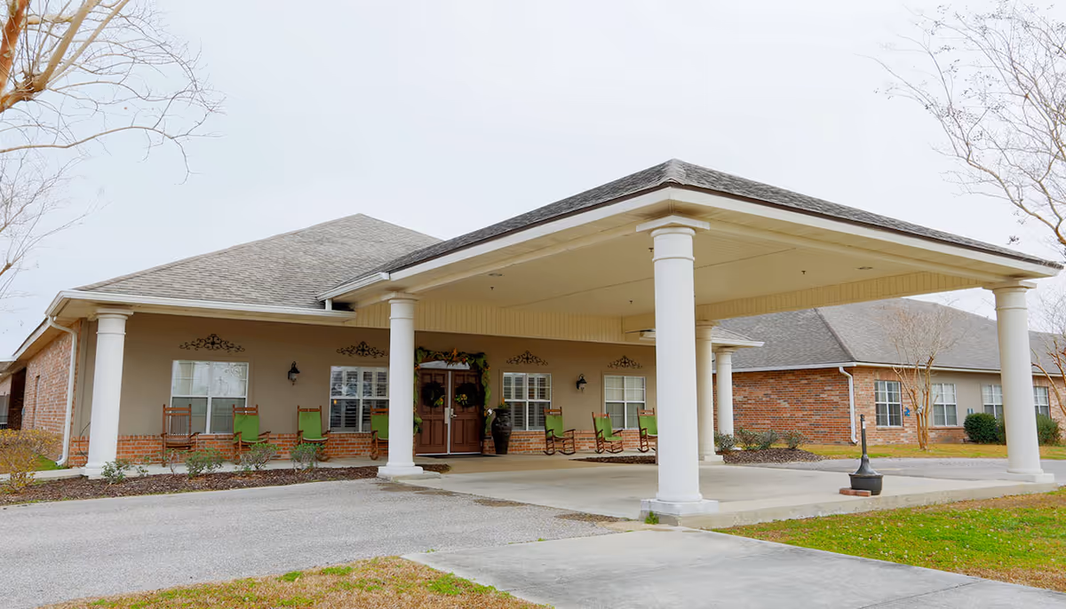 Front entrance of a single-story senior living building with a covered porte-cochere, white columns, and rocking chairs on the porch.