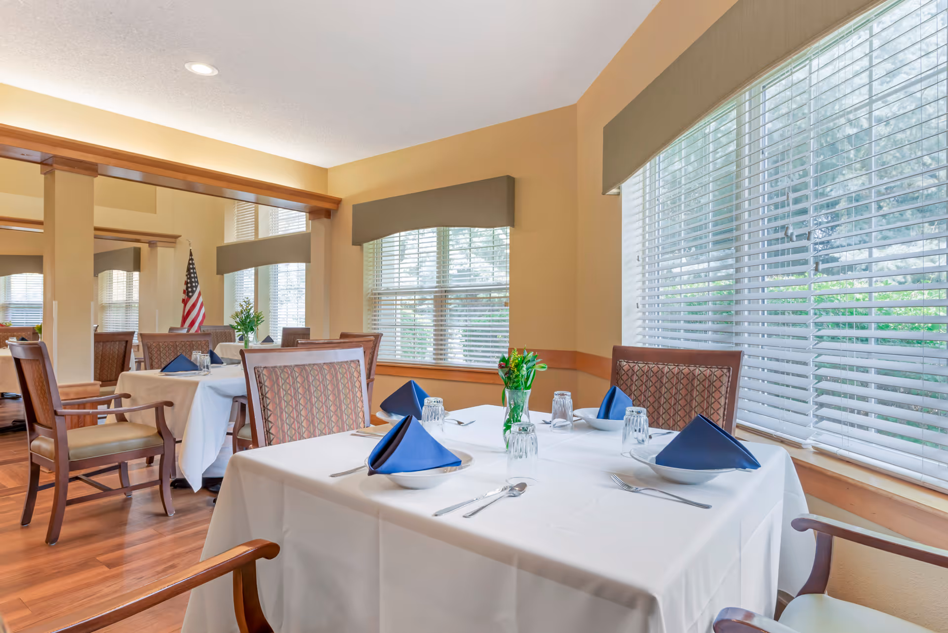 A bright dining room with tables covered in white tablecloths, set with blue folded napkins, plates, utensils, and glasses. The room has large windows with blinds, wooden chairs with patterned upholstery, and a vase with flowers on each table. An American flag is visible in the background.