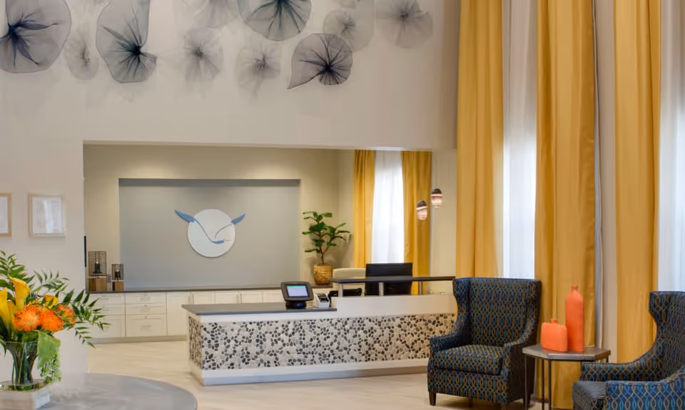 Reception area of Beach House Assisted Living & Memory Care with a front desk featuring a pebble-patterned front, a computer, and a logo on the wall behind. Two patterned armchairs and a small table with decorative orange vases are positioned near tall windows with yellow curtains. A plant and wall art with translucent, abstract shapes are also visible.