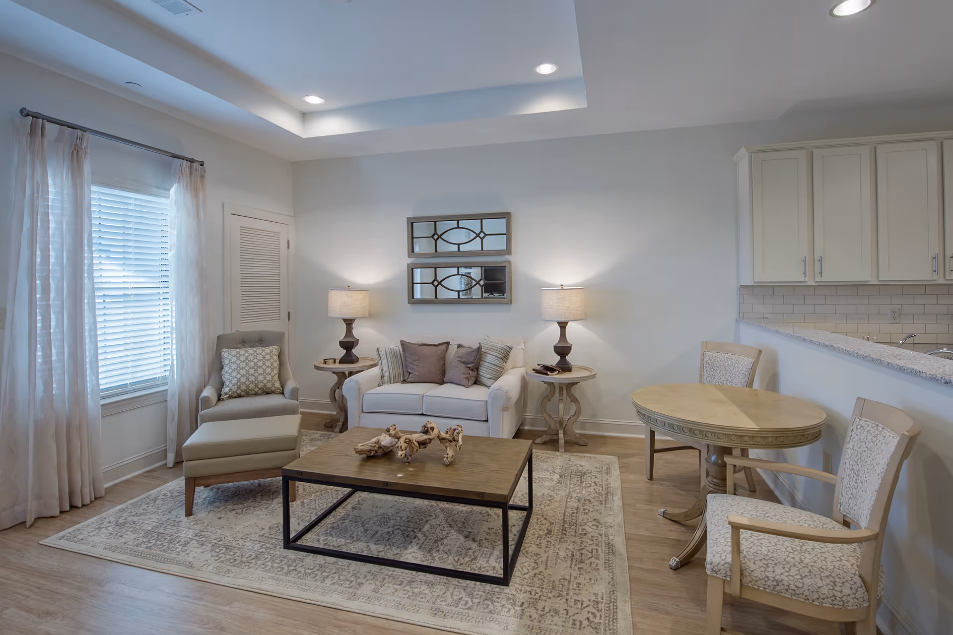 A cozy living room with a white loveseat adorned with multiple pillows, flanked by two side tables each holding a lamp. To the left, there is a cushioned armchair with an ottoman near a window with sheer curtains. In front of the loveseat is a wooden coffee table with decorative pieces on a patterned area rug. To the right, there is a small round dining table with two upholstered chairs. The kitchen area with white cabinets and a tiled backsplash is partially visible in the background.