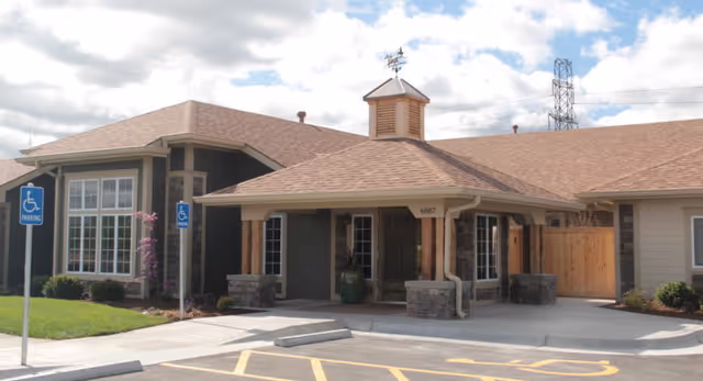 Exterior view of a single-story building with a covered entrance supported by wooden pillars. The building has a brown shingled roof and beige siding. There are two handicap parking signs and spaces in front of the entrance. The sky is partly cloudy.