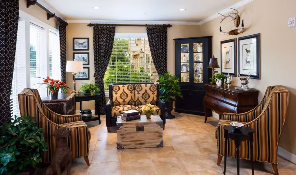 Bright, well-decorated living room with patterned armchairs and a loveseat arranged around a wooden trunk coffee table, large windows with drapes, cabinets, and decorative accents.