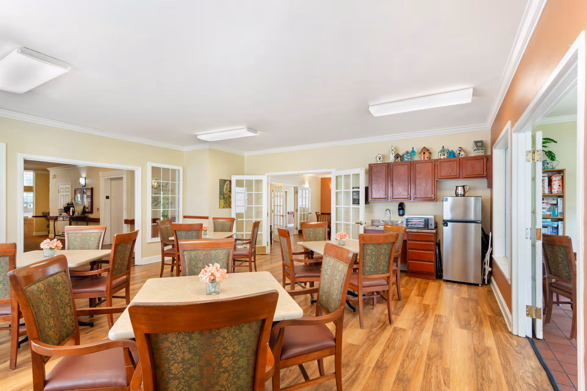 A bright dining room in a senior living facility with multiple square tables surrounded by wooden chairs with patterned upholstery. Each table has a small vase with pink flowers. The room has light-colored walls, wooden flooring, and a small kitchenette area with a stainless steel refrigerator, cabinets, and a countertop. French doors and windows open into adjacent rooms.