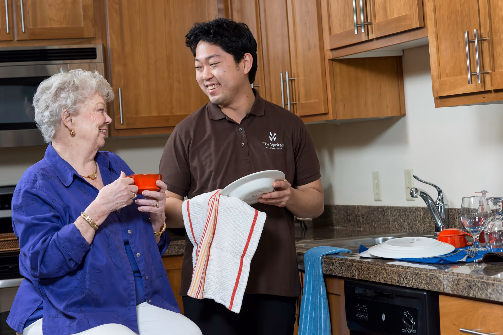 An elderly woman holding a red cup and smiling at a younger man who is drying a white plate with a dish towel in a kitchen with wooden cabinets and granite countertops.