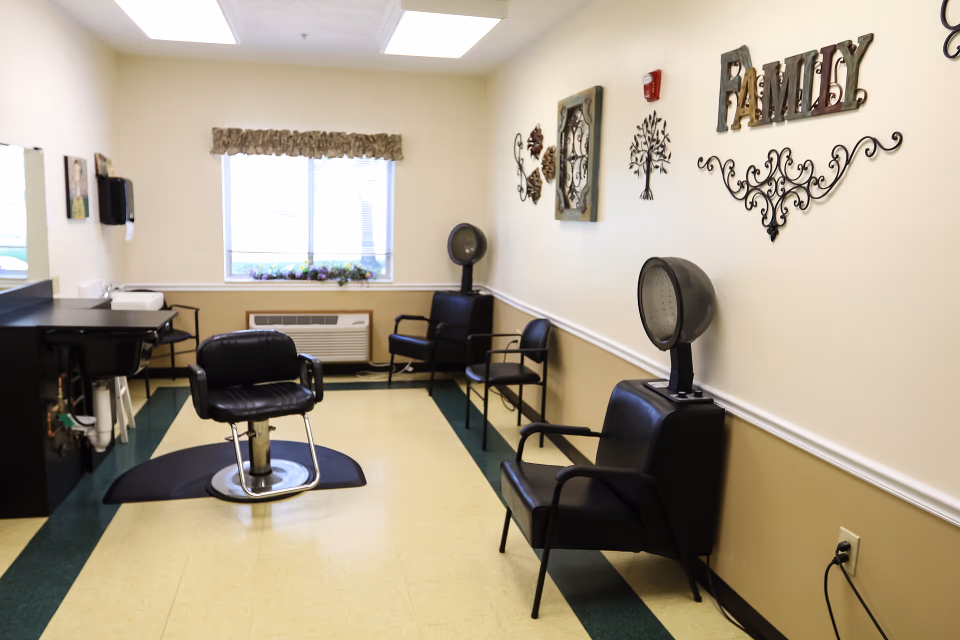 Interior view of a hair salon area in a senior living facility with black salon chairs, hair dryers, a sink, and wall decorations including the word 'FAMILY'.