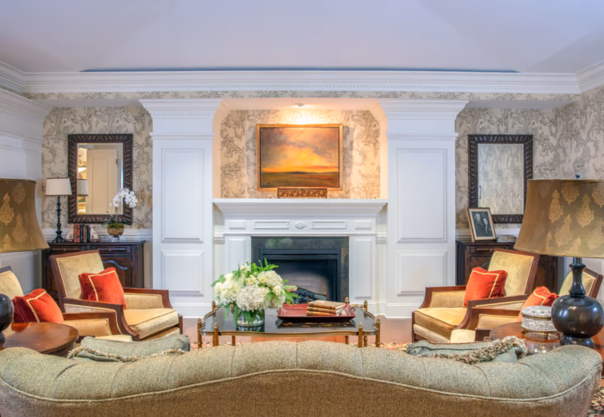 A cozy living room with a beige sofa and two armchairs with red cushions facing a white fireplace. Above the fireplace is a painting of a sunset. The room features floral wallpaper, two large mirrors on either side of the fireplace, wooden side tables with lamps, and a coffee table with a vase of white flowers and some books.