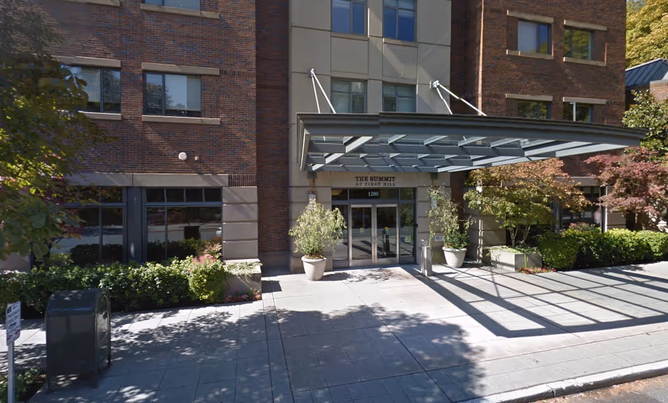 Street-level view of a brick senior living building entrance with a glass canopy, potted plants, and double doors.