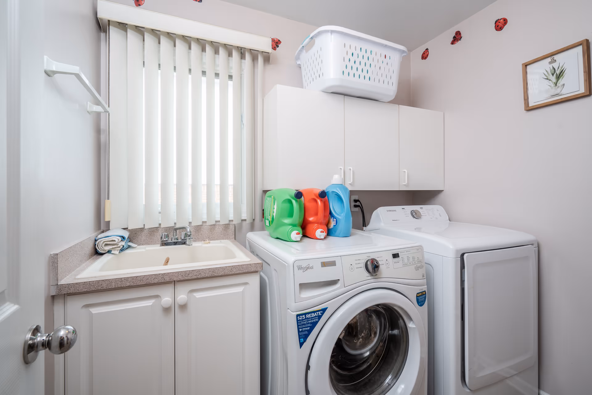 A laundry room with a front-loading washing machine and a top-loading dryer side by side. On top of the washing machine are three large detergent bottles in green, red, and blue. Above the machines, there are white cabinets with a white laundry basket on top. To the left, there is a countertop with a sink and a window with vertical blinds. The walls are decorated with small ladybug stickers and a framed picture of a plant.