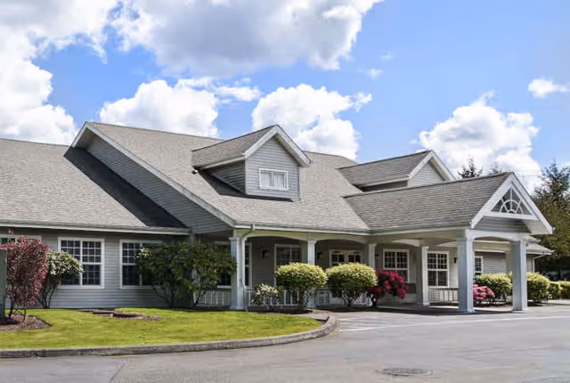 Front exterior of a single-story senior living building with a covered entrance, pitched roof, and landscaped shrubs under a partly cloudy sky.