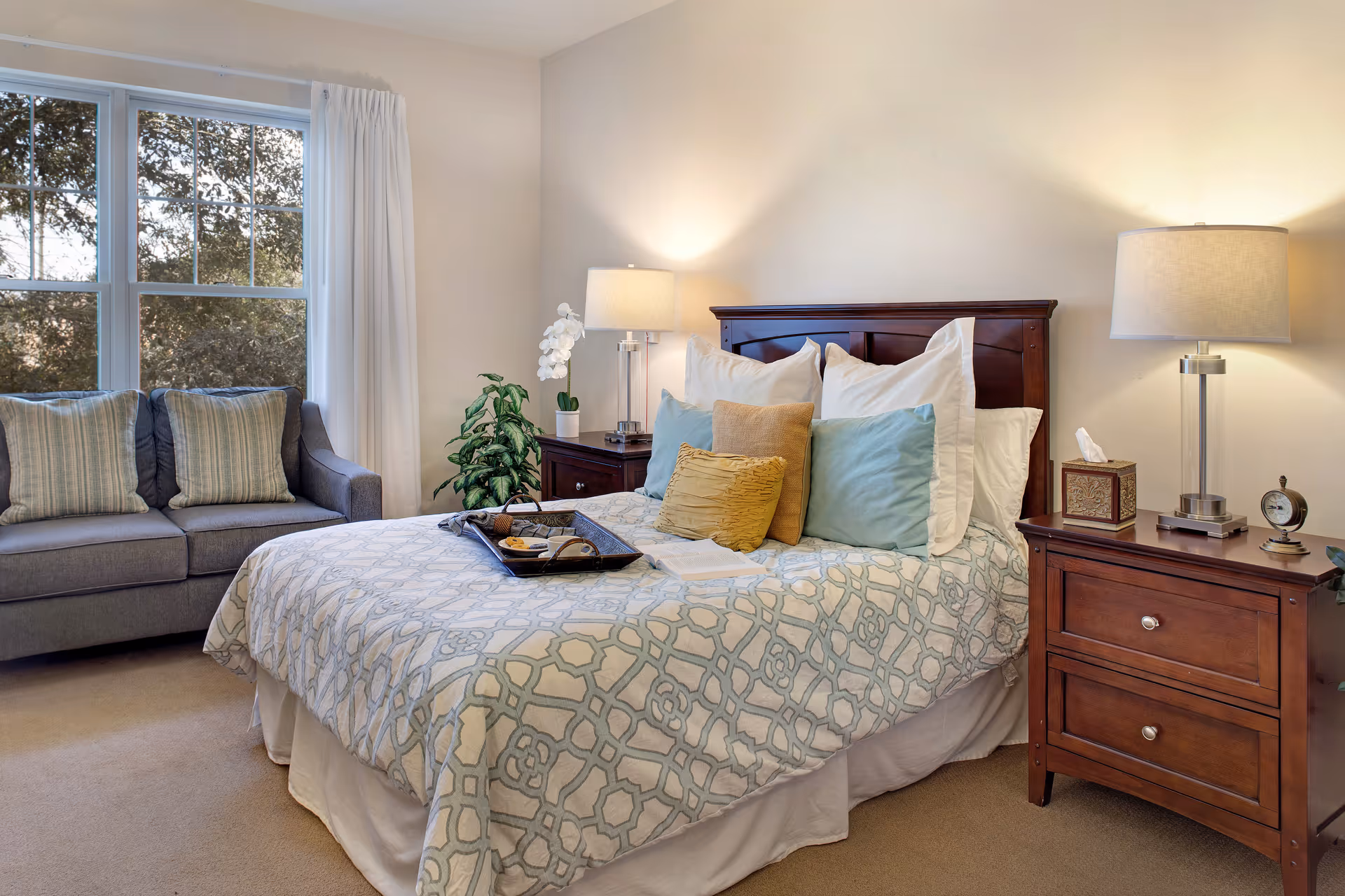 A cozy bedroom with a neatly made bed featuring patterned bedding and multiple pillows. There are two wooden nightstands on either side of the bed, each with a modern lamp. A gray sofa with striped cushions is positioned near a large window with white curtains, and there are green plants adding a touch of nature to the room.