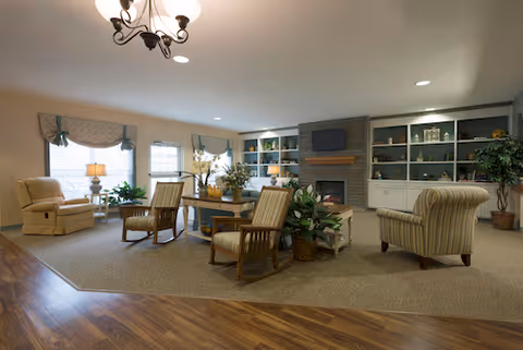 Spacious common living room with upholstered armchairs and wooden chairs arranged around a central table, a fireplace flanked by built-in shelves, potted plants, and large windows with valances.