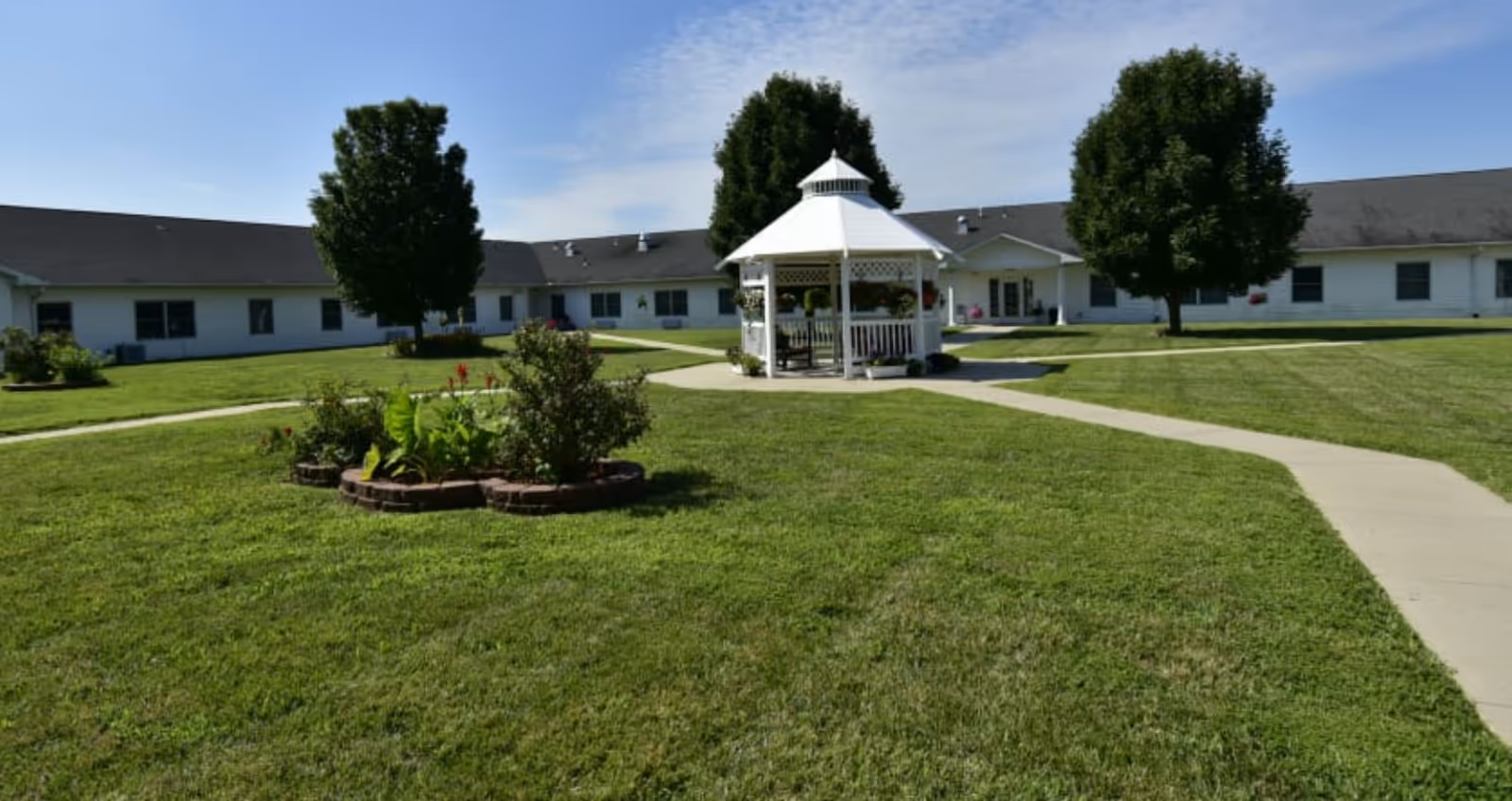 A well-maintained outdoor courtyard area with a white gazebo in the center, surrounded by green grass, a flower bed, and several trees. A paved walkway leads to the gazebo and extends around the courtyard, with a single-story building in the background under a clear blue sky.
