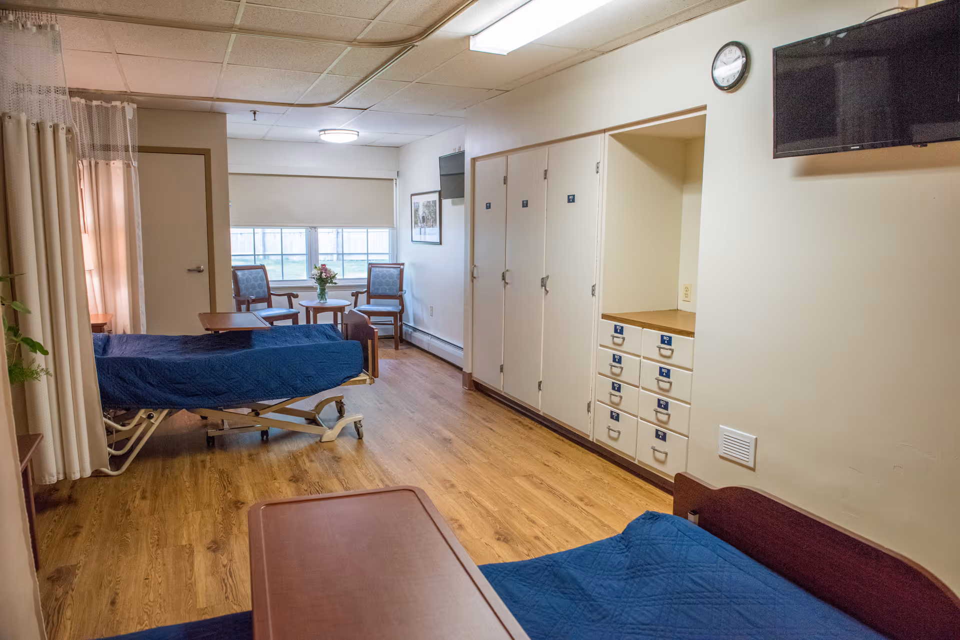 A senior living facility room with two hospital-style beds covered in blue blankets, a wooden floor, a wall-mounted TV, a clock, and a seating area with two chairs and a small table near a window with blinds.