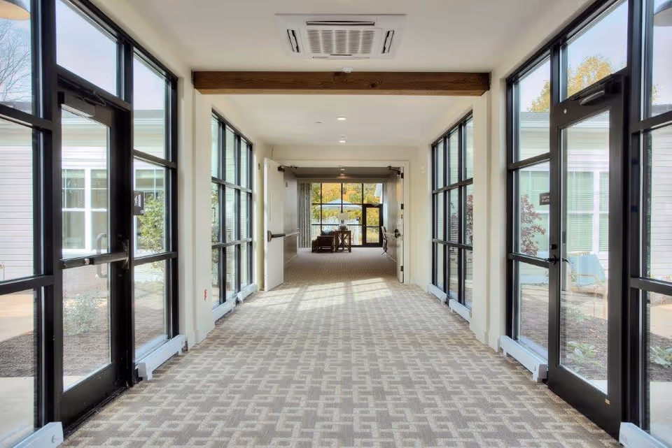 A bright hallway with large glass doors and windows on both sides, leading to a common area with seating and a view of an outdoor space with trees and a fence.