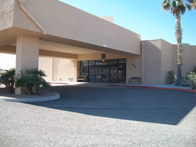 Entrance of a beige stucco building with a covered drop-off area, glass double doors, two benches on either side of the entrance, and palm trees nearby under a clear blue sky.