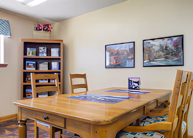 A cozy dining room with a wooden table and four wooden chairs with patterned cushions. On the table are placemats and a small decorative item. In the background, there is a wooden bookshelf with various items and two framed paintings on the beige wall.
