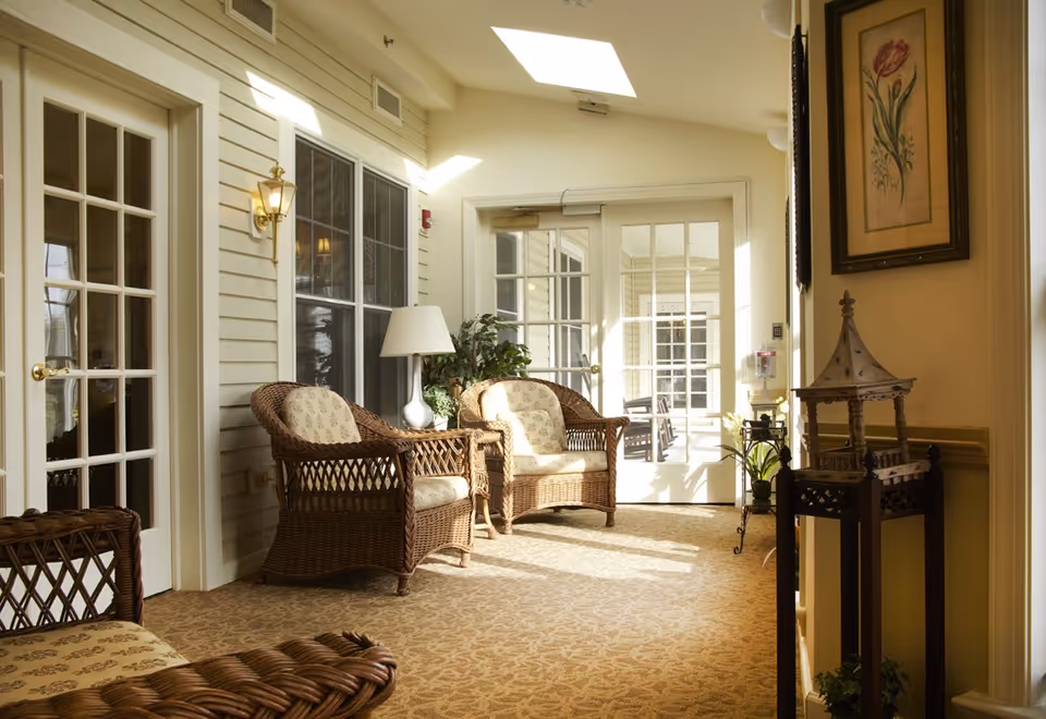 Sunlit sitting area with wicker chairs, a side table and lamp beside French doors.