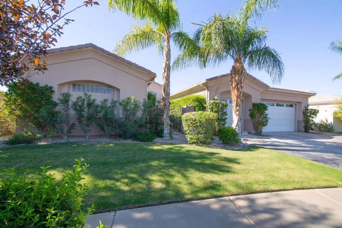 Exterior view of a single-story residential building with a beige stucco finish, a tiled roof, and a white garage door. The front yard features a well-maintained green lawn, various shrubs, and two tall palm trees. A paved driveway and sidewalk are visible in the foreground under a clear blue sky.