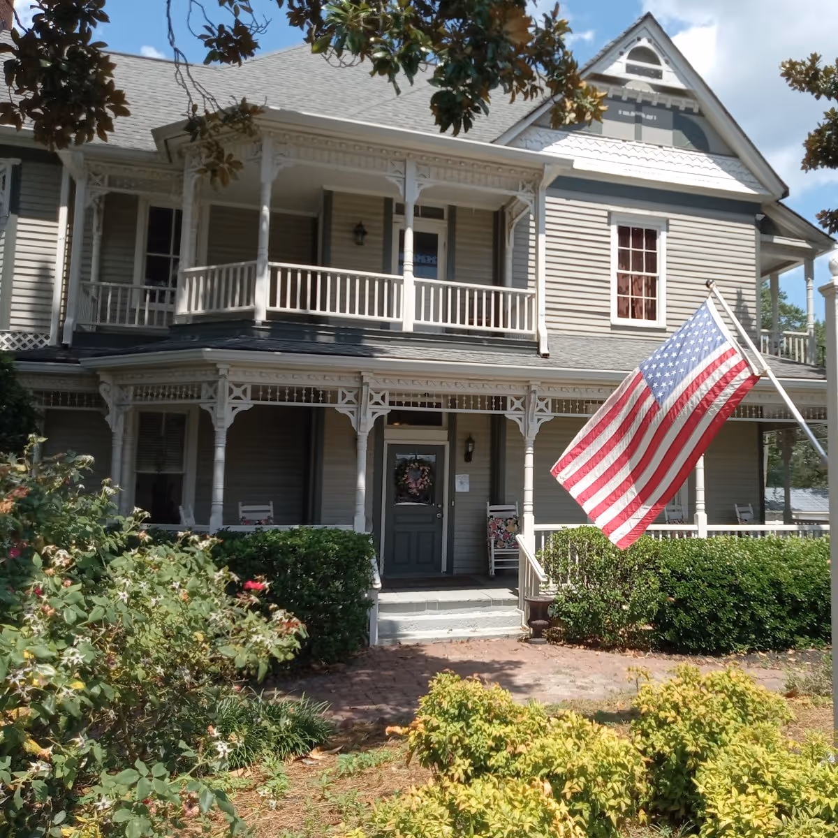 Victorian-style two-story house with a covered front porch, balcony, and an American flag displayed by the entrance.