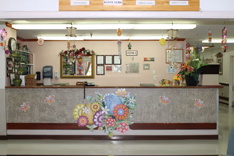 Reception desk area decorated with colorful flower and butterfly decals. Behind the desk are framed certificates, a mirror, and a sign labeled 'STATION A'. The desk has a telephone and a large flower arrangement on the right side. The ceiling has fluorescent lights and hanging decorations.