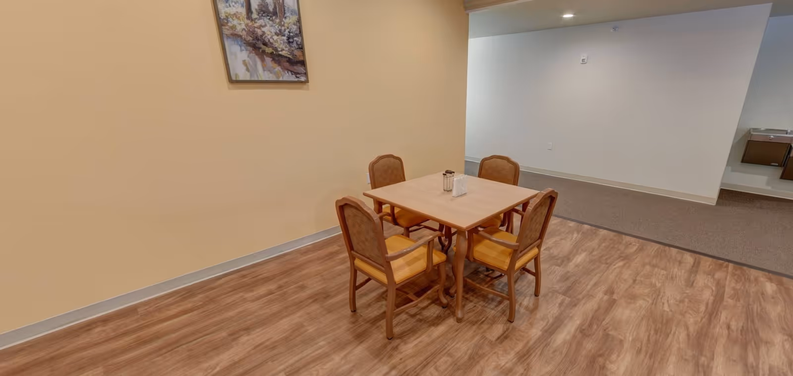 A small dining area with a square wooden table and four wooden chairs with yellow cushions. The room has light brown wooden flooring, beige and white walls, a painting on the beige wall, and a water fountain mounted on the white wall in the background.