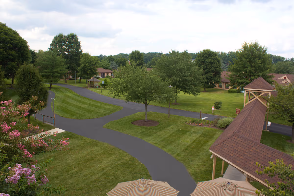 Landscaped outdoor grounds with paved paths, lawns, trees, a covered wooden walkway and patio umbrellas at a senior living village.