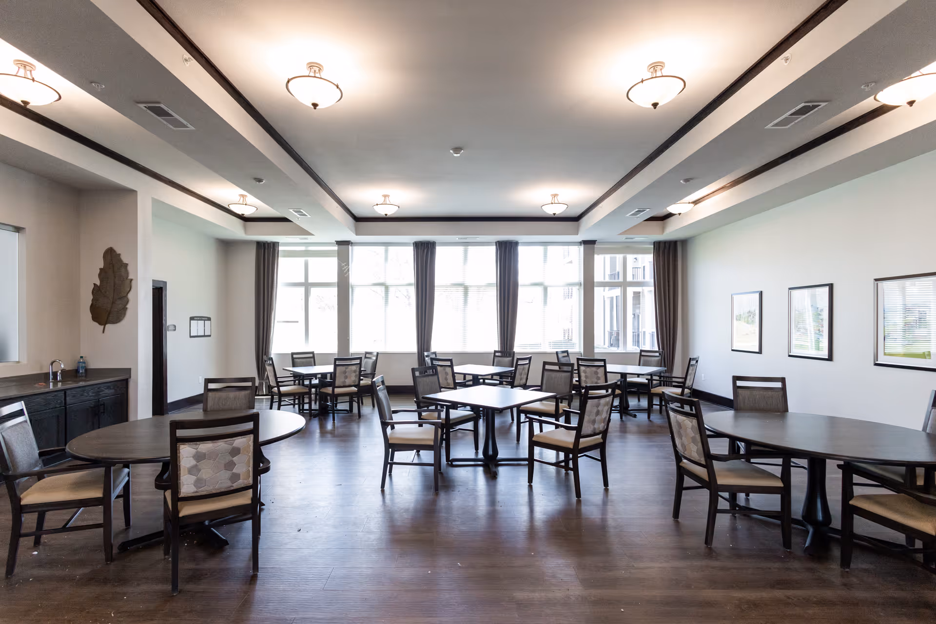 A spacious dining room with multiple round and square tables surrounded by chairs. The room has large windows with curtains allowing natural light to fill the space. The ceiling features several light fixtures, and the floor is made of dark wood. There are framed pictures on one wall and a decorative leaf sculpture on another wall near a countertop with a sink.
