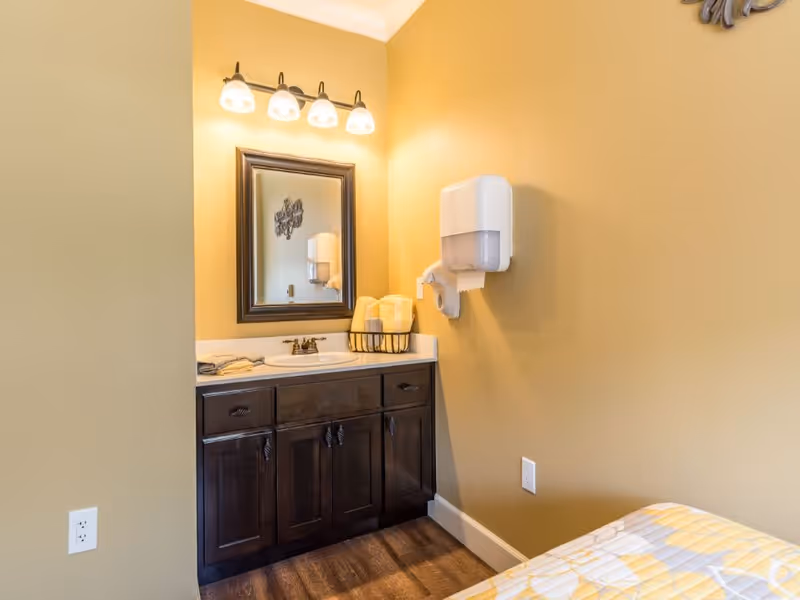 Small vanity with sink and mirror in a warm yellow room, a wall-mounted paper dispenser and towels, with part of a bed visible.