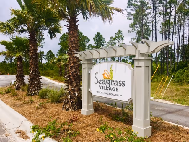 Outdoor view of a sign for Seagrass Village Senior Living Community, mounted on a white pergola-style frame, surrounded by palm trees and landscaping along a paved road.