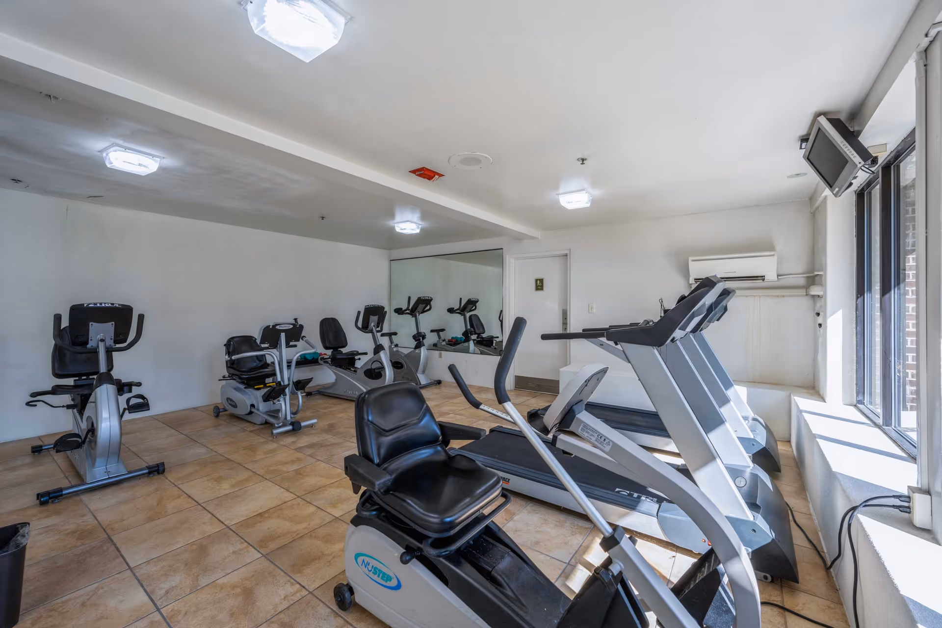 A small fitness room with exercise equipment including stationary bikes, recumbent bikes, and treadmills. The room has tiled floors, white walls, a large mirror on one wall, and windows letting in natural light. A wall-mounted TV is positioned near the windows.