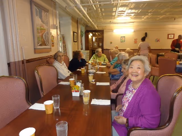 A group of elderly women sitting around a long wooden dining table in a well-lit room. The women are engaged in conversation and appear to be enjoying each other's company. The room has pink cushioned chairs, framed artwork on the walls, and a decorative ceiling. There are cups, glasses, and napkins on the table.