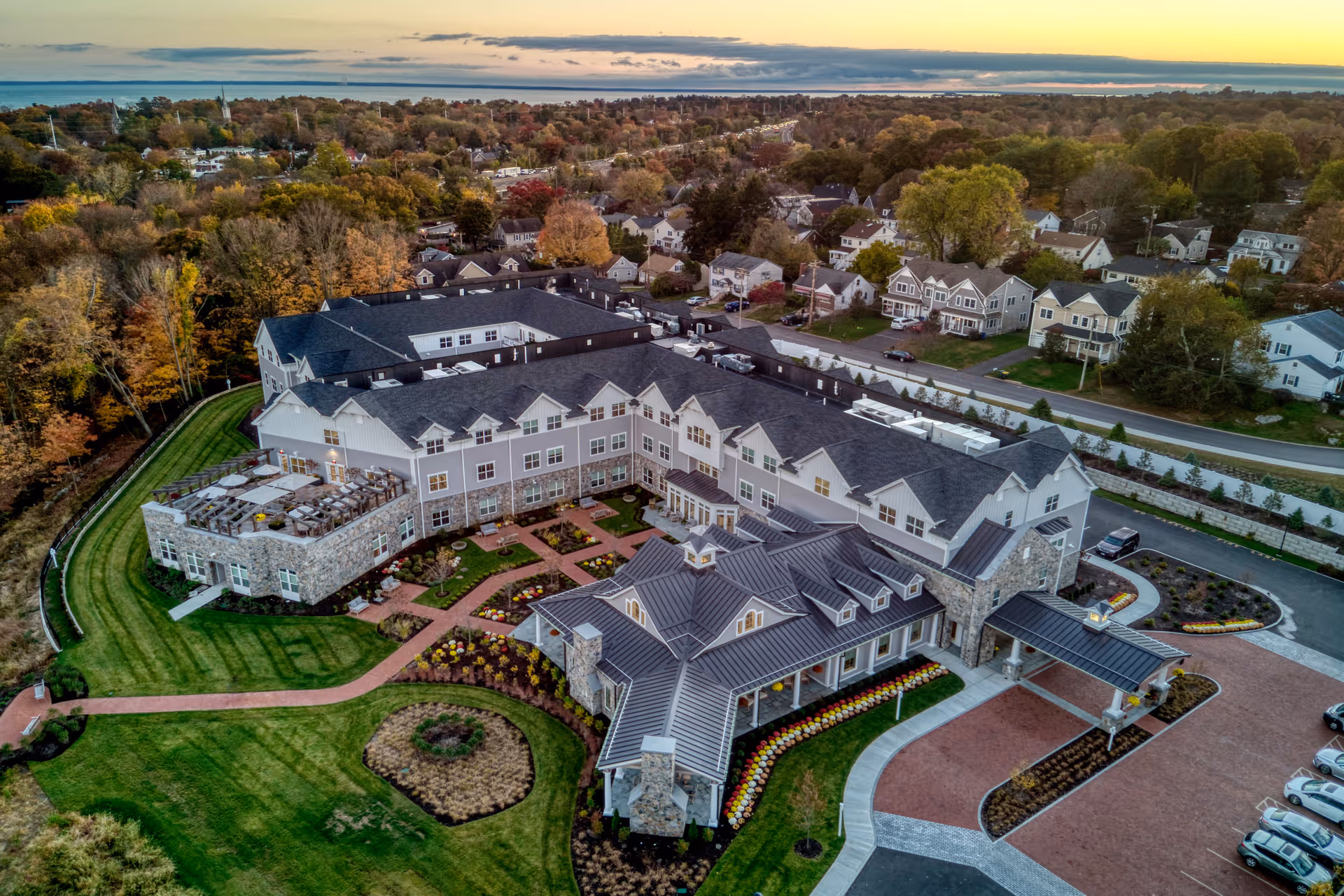 Aerial view of Maplewood at Southport senior living facility during sunset, showing a large multi-wing building with stone and siding exterior, landscaped gardens, outdoor seating area, and surrounding residential neighborhood.
