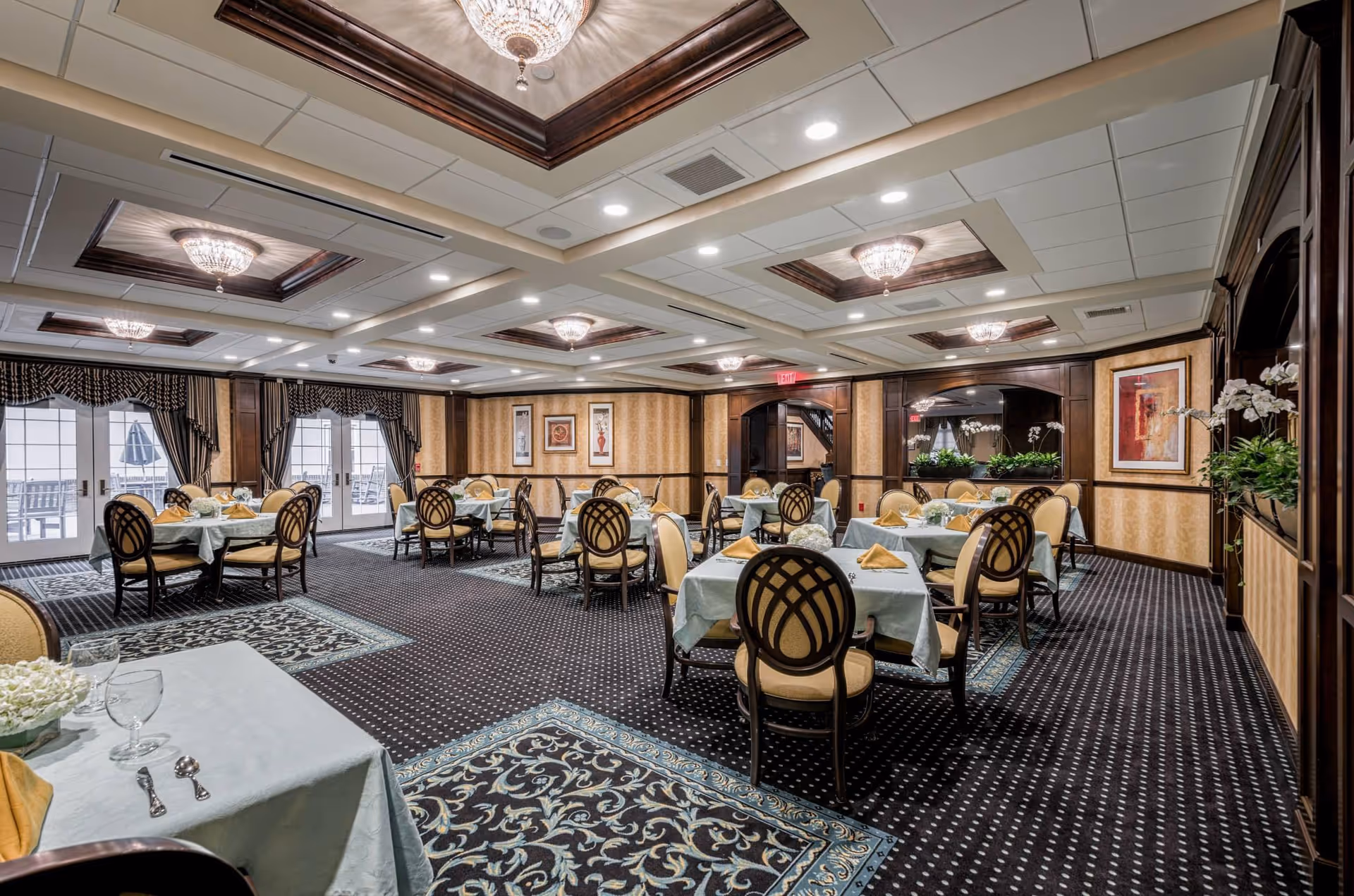 Elegant dining room with multiple round tables covered with light blue tablecloths and set with yellow napkins, glassware, and silverware. The room features patterned carpet, ornate ceiling light fixtures, large windows with draped curtains, and framed artwork on the walls.