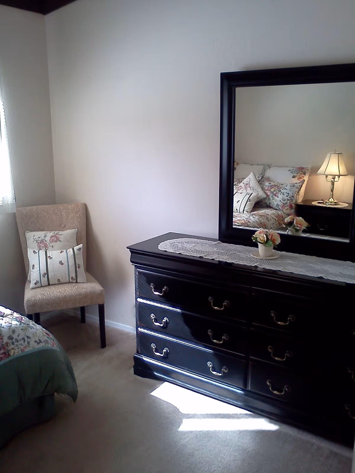A bedroom corner featuring a dark wooden dresser with a large mirror on top, decorated with a lace runner and a small flower arrangement. To the left, there is a beige upholstered chair with two floral-patterned pillows. Part of a bed with floral bedding is visible on the left side of the image.