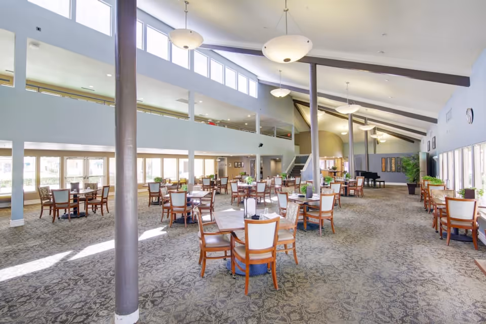 A spacious, well-lit dining area with multiple wooden tables and chairs arranged neatly on a patterned carpet. Large windows along the walls allow natural light to fill the room. The ceiling is high with modern light fixtures hanging down, and there is a staircase leading to an upper level. Potted plants and a piano are visible in the background.