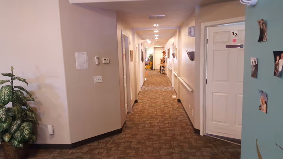 A long, carpeted hallway in an assisted living facility with beige walls and several doors on both sides. There is a potted plant on the left side near the wall, and some photos are attached to the right wall near a door labeled '12'. The hallway is well-lit with ceiling lights and has handrails along the walls.