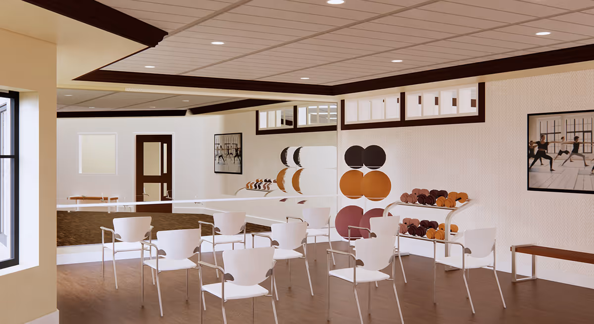 A fitness or exercise room with white chairs arranged in rows facing a mirrored wall. The room has wooden flooring, a rack with various colored exercise balls, and a shelf with dumbbells. There are framed pictures on the walls showing people doing yoga or stretching exercises. The ceiling has recessed lighting and dark wood trim.
