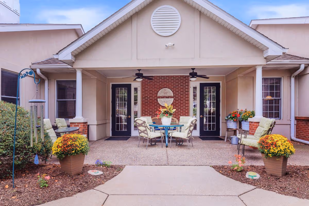 Outdoor covered patio area at Country Manor Memory Care with a round glass table surrounded by four cushioned chairs. Two ceiling fans are mounted above. There are two black doors with glass panes on a brick wall, flanked by beige walls and windows. Potted yellow and orange flowers and green shrubs decorate the area, along with a wind chime on the left side.