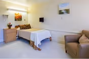 A clean, simply furnished patient bedroom with a single bed, nightstand, armchair, wall-mounted TV, and artwork on the walls.