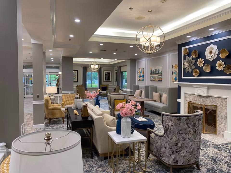 A spacious and elegantly decorated living room area in a senior living facility. The room features multiple seating options including armchairs, sofas, and built-in bench seating with cushions. There are decorative vases with pink flowers on tables, a fireplace with floral wall art above it, and modern light fixtures hanging from the ceiling. Large windows allow natural light to enter the room.