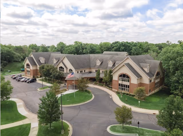 Aerial view of a large two-story senior living building with a circular driveway, flagpole, parking lot, and surrounding trees.